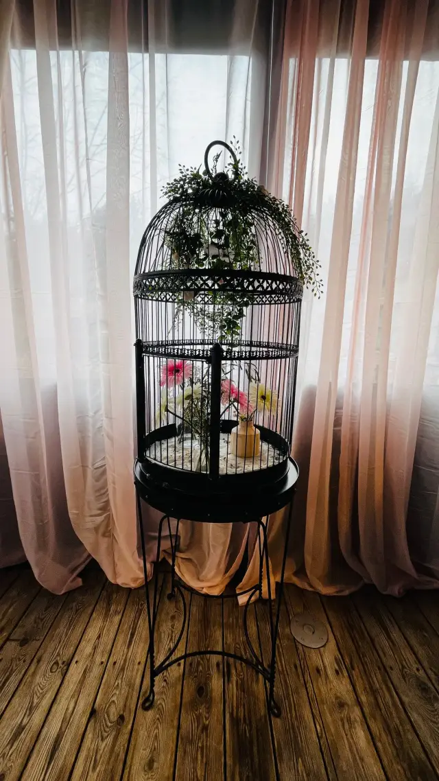 Decoratieve vogelkooi met kunstbloemen op een sierlijk onderstel voor een raam met roze en witte gordijnen
