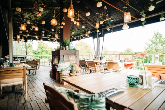 Overdekt terras met houten tafels en stoelen, hangende lampen en rozen aan het plafond, uitzicht op groen